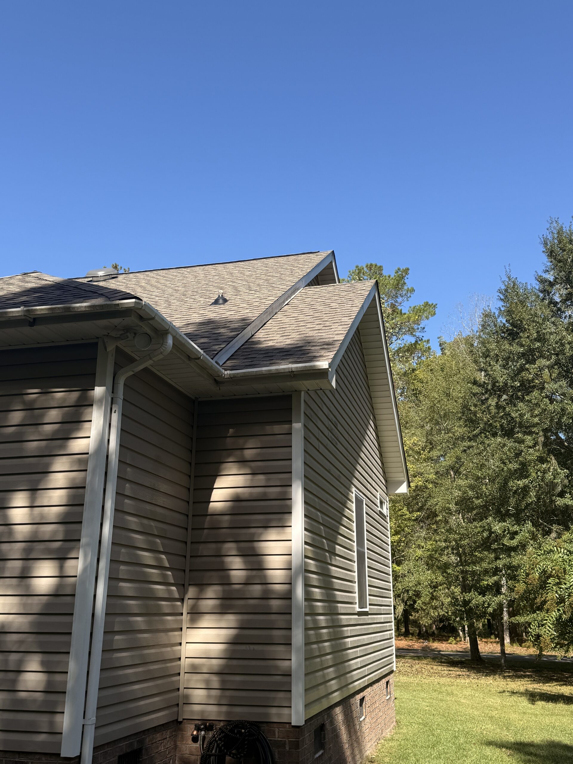 A tan house with vinyl siding and a shingled roof is shown next to a grassy yard and trees under a clear blue sky.