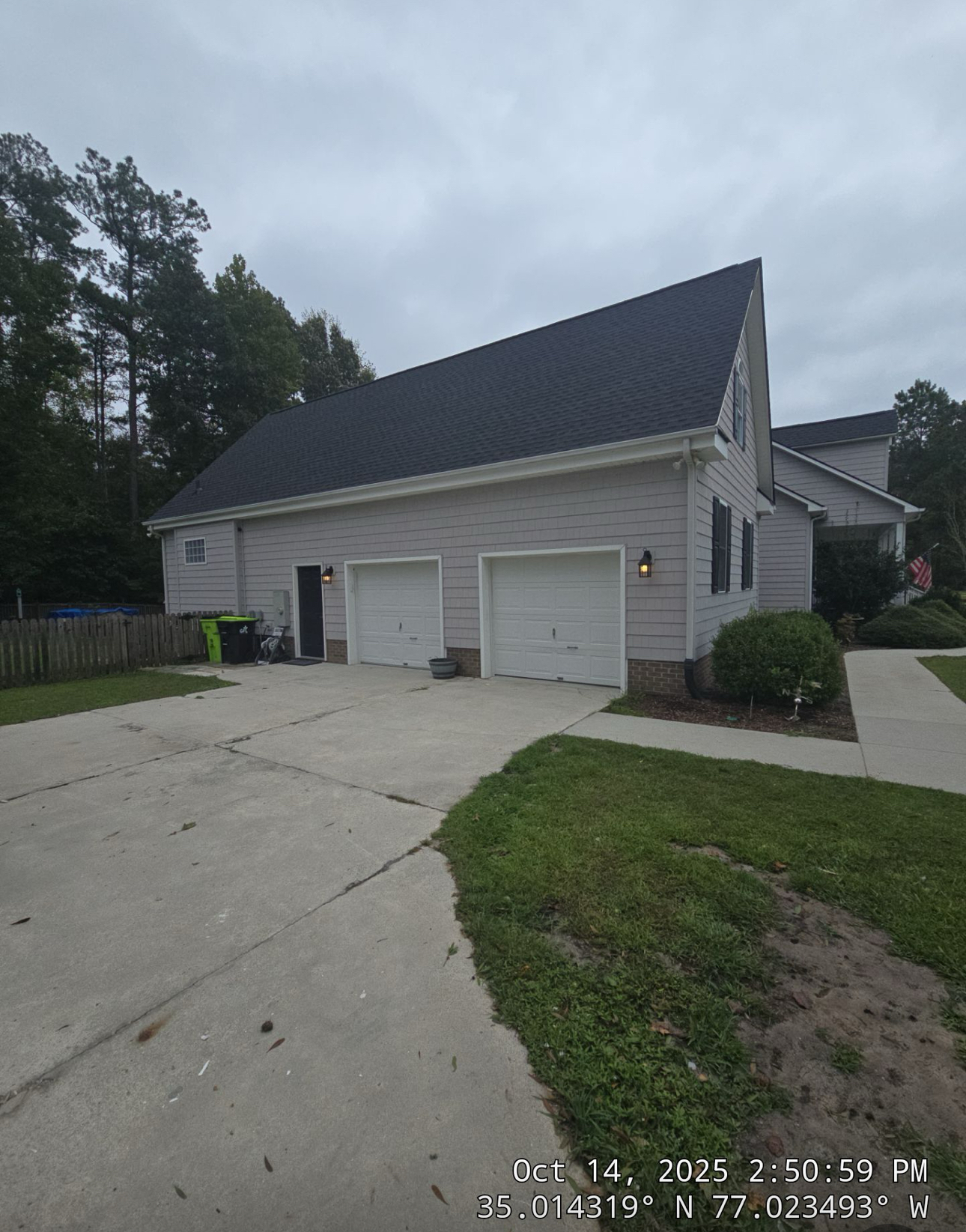 A two-car garage attached to a light gray house with a sloped roof, concrete driveway, grass, and timestamp with coordinates in the lower right corner.