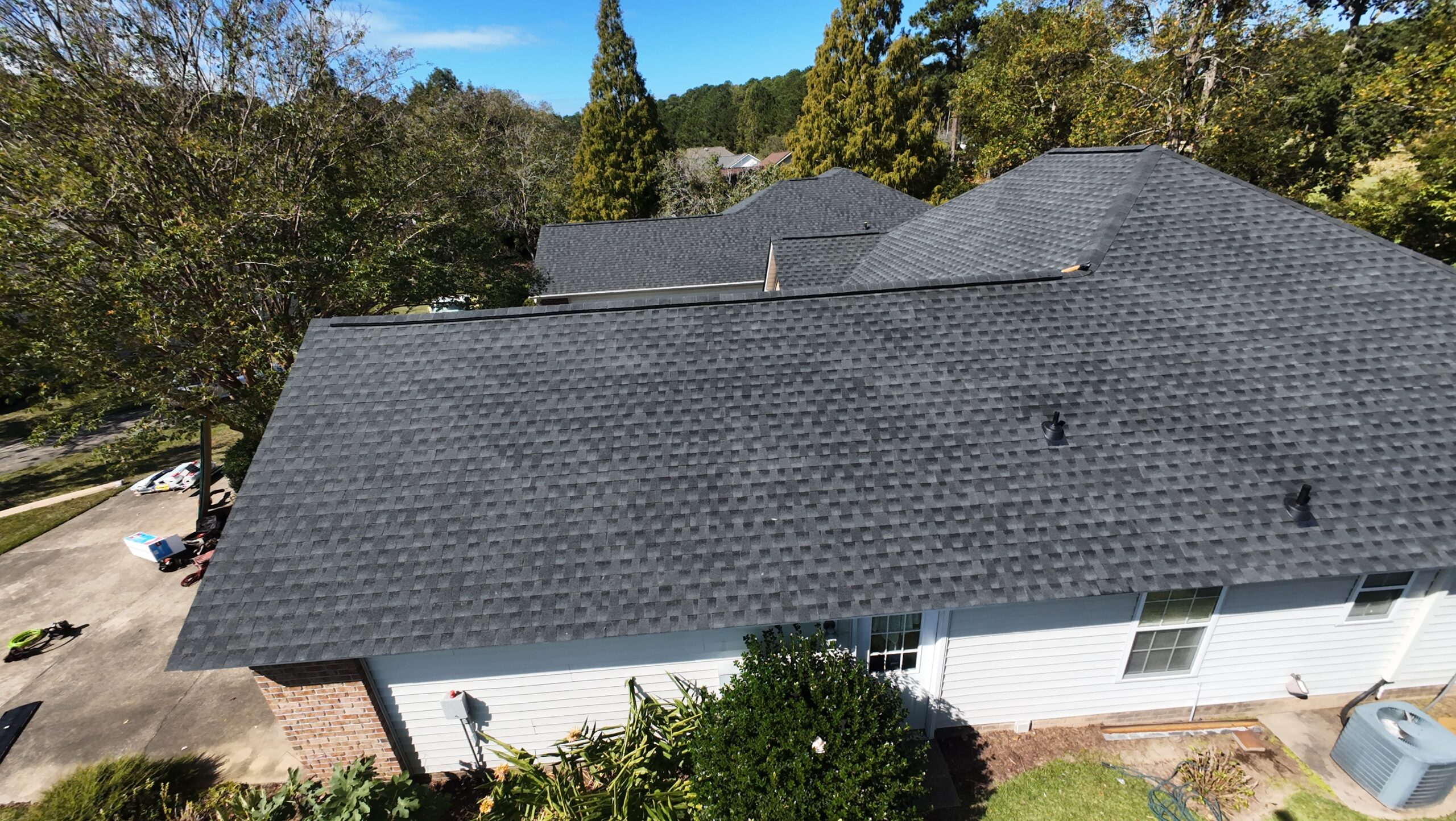 Aerial view of a house with a new dark gray shingle roof, surrounded by trees and a driveway, on a sunny day.