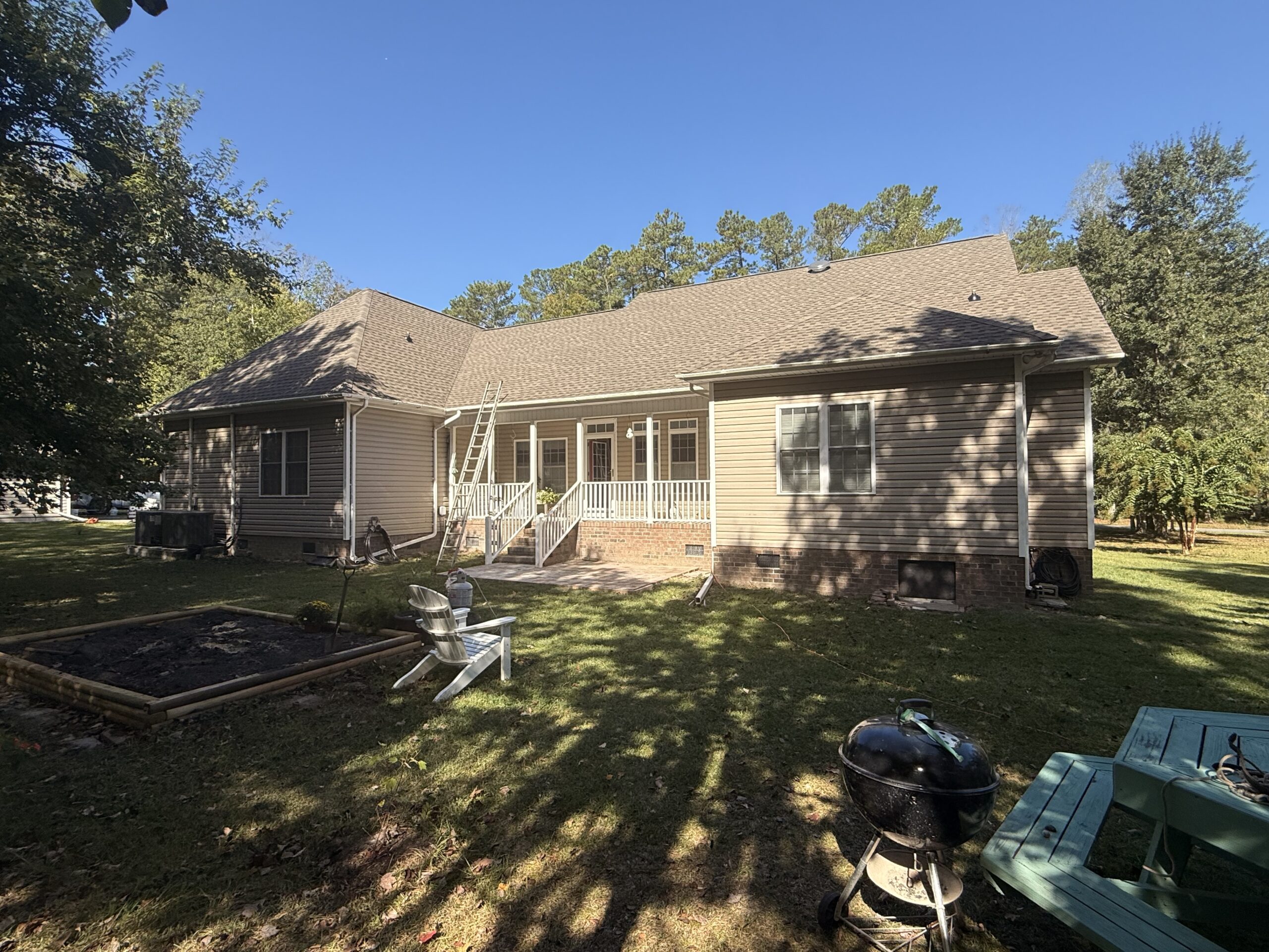A single-story house with beige siding, a small back porch, and a ladder leaning against the roof. There are lawn chairs, a grill, and a picnic table in the backyard.