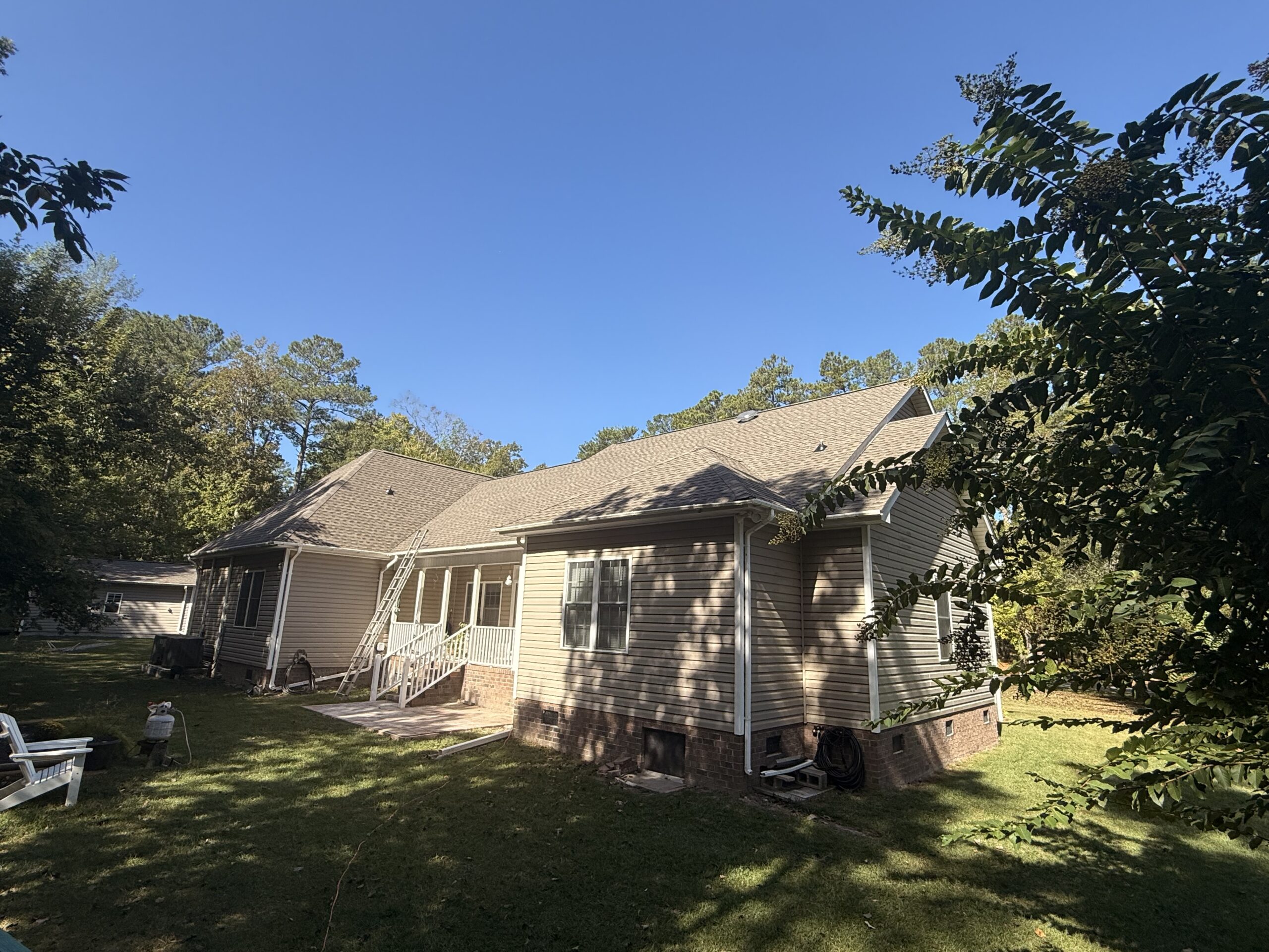 Single-story beige house with a gable roof, white trim, and two small porches; surrounded by trees and a grassy yard under a clear blue sky.