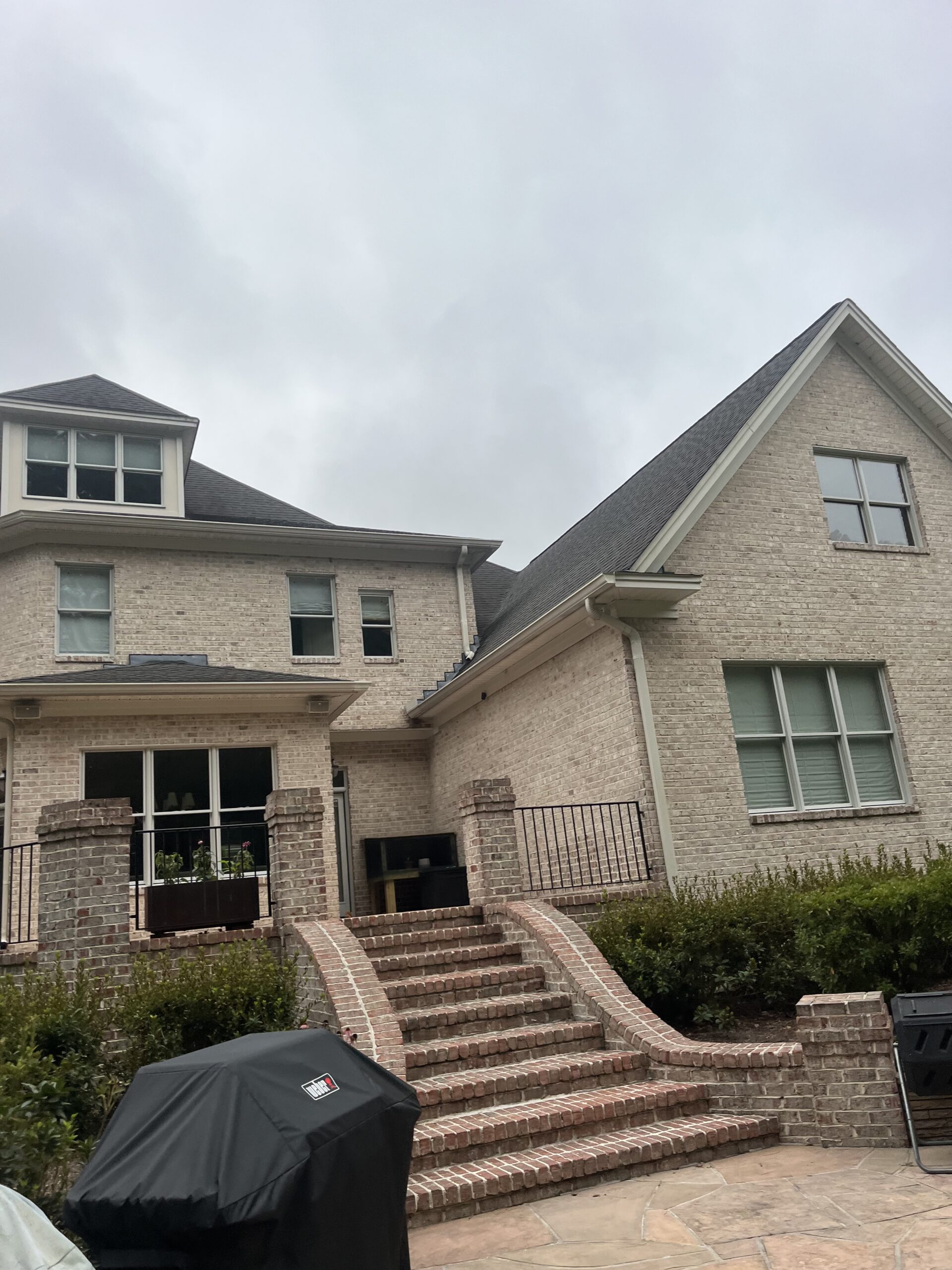 Two-story beige brick house with multiple windows, a sloped dark roof, and a brick staircase leading to a raised patio. Shrubs line the front and a barbecue grill is in the foreground.